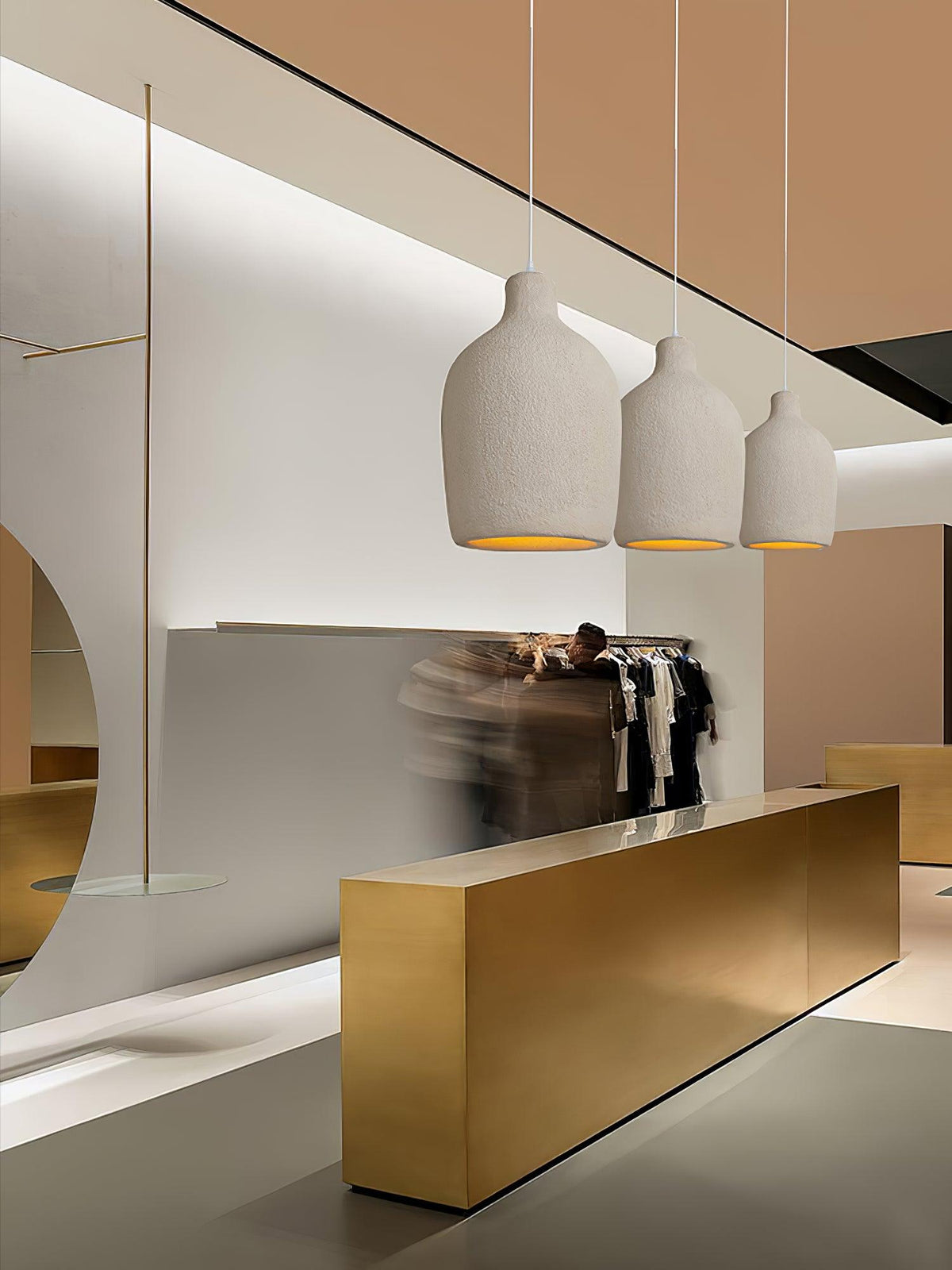 A row of three beige Clérachic pendant lights in graduated sizes, seen over a gold counter and a rail of clothes.