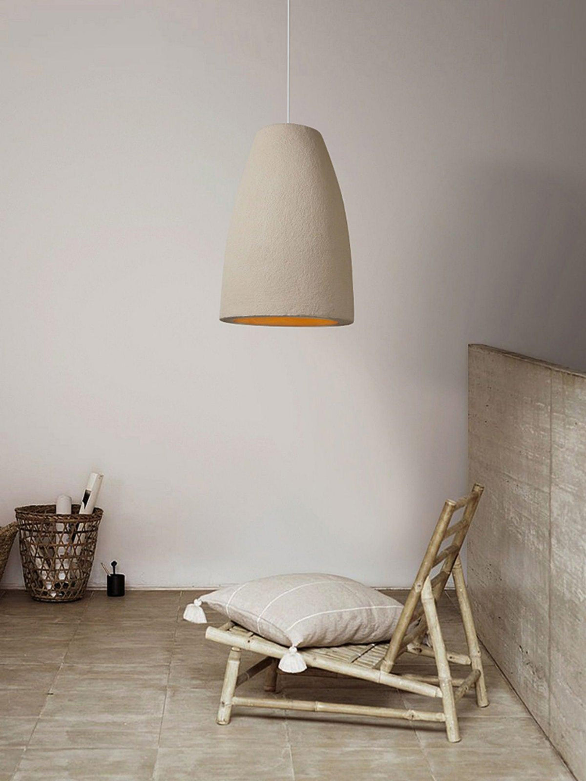 A tapered beige Clérachic pendant light on a pale wall, with a woven basket and a bamboo chair with a cushion below.