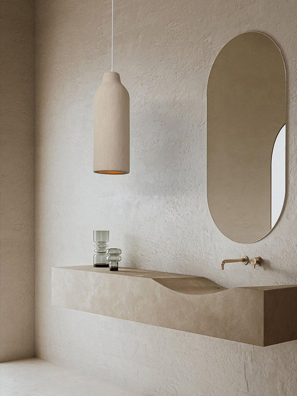 Narrow beige Clérachic pendant light beside an oval mirror, above a pale stone washbasin with a brass tap.