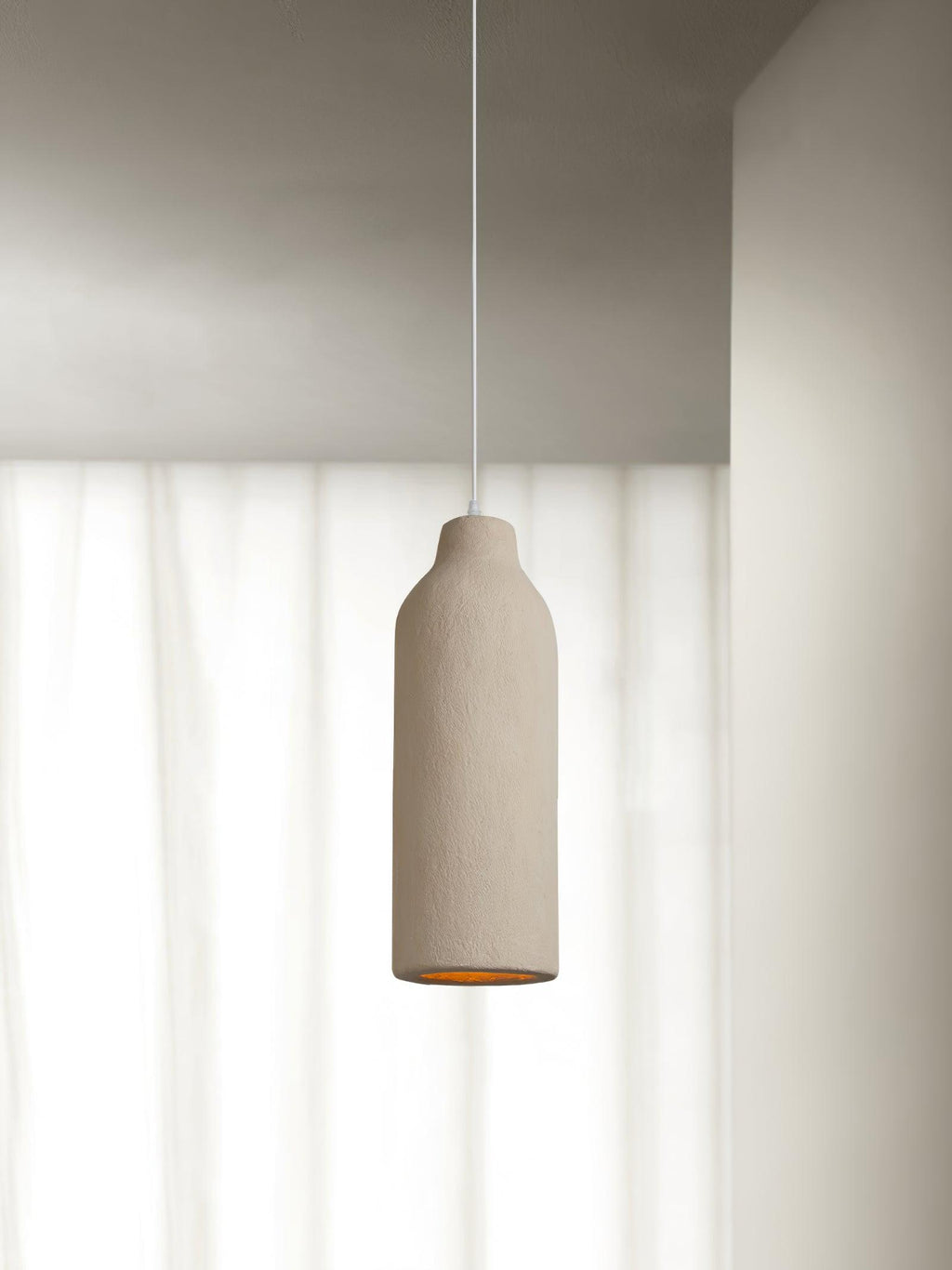 A tall narrow beige Clérachic pendant light in front of sheer white curtains, with its amber opening just visible below.