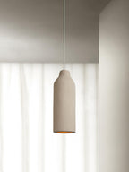 A tall narrow beige Clérachic pendant light in front of sheer white curtains, with its amber opening just visible below.