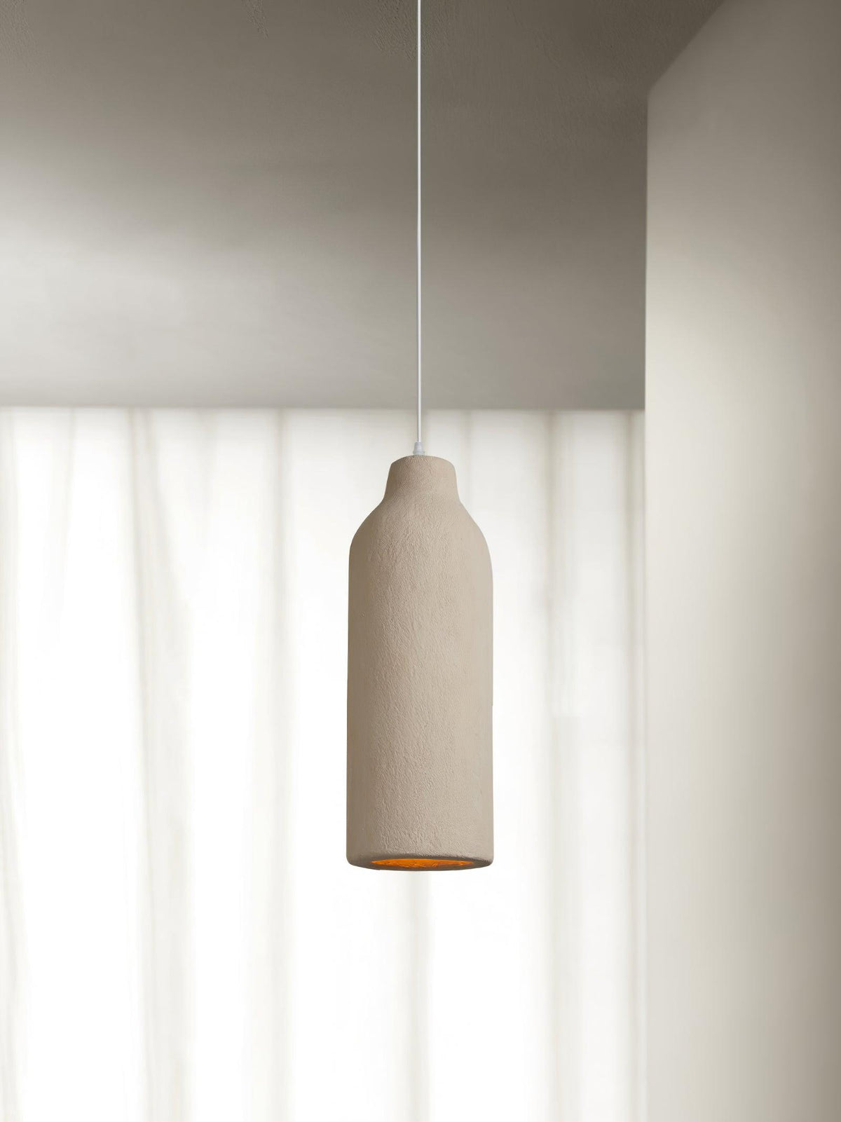 A tall narrow beige Clérachic pendant light in front of sheer white curtains, with its amber opening just visible below.