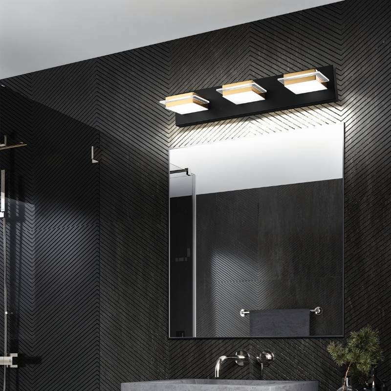 Clérachic three-head black bar light with gold bands and frosted square diffusers, mounted above a mirror in a dark chevron-tiled bathroom.