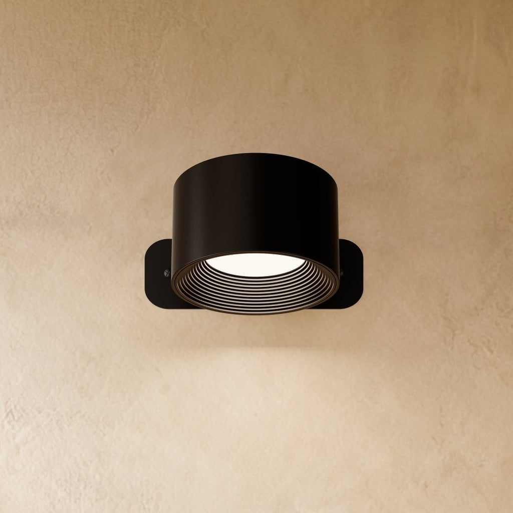 Clérachic compact black round wall lamp with a short drum head, lit, its ribbed inner ring glowing white.