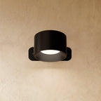 Clérachic compact black round wall lamp with a short drum head, lit, its ribbed inner ring glowing white.