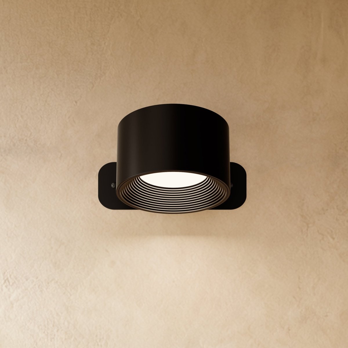 Clérachic compact black round wall lamp with a short drum head, lit, its ribbed inner ring glowing white.