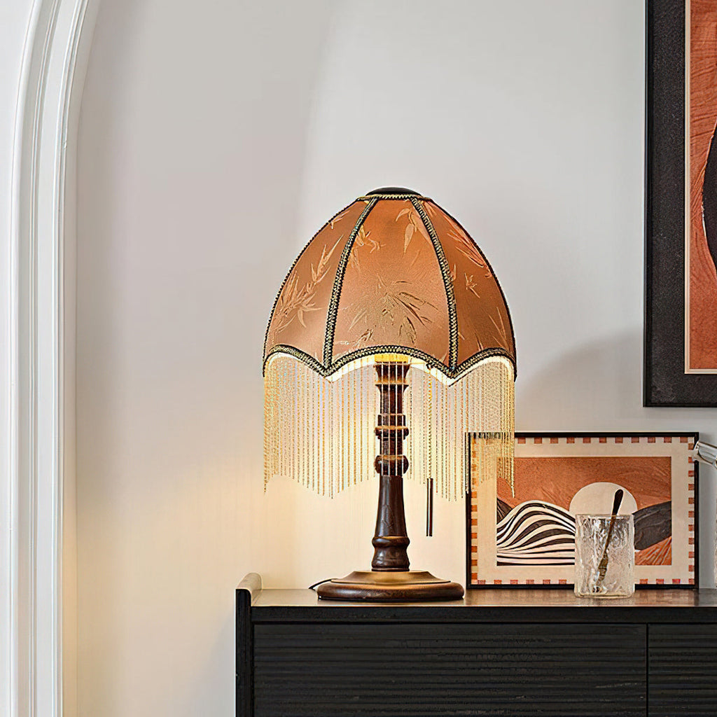 Warm light shines through the brown shade of this Clérachic lamp, its beaded fringe hanging above a sideboard and a framed abstract landscape.