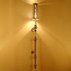 Clérachic slim beaded wall lamp with a tapered clear shade, glowing warm and casting light up a golden wall.