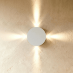 Clérachic white round wall lamp with four narrow beams of warm light fanning out across a plastered wall.