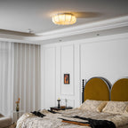 A small, lit Clérachic fabric ceiling light seen from the far side of a white panelled bedroom with a mustard headboard.