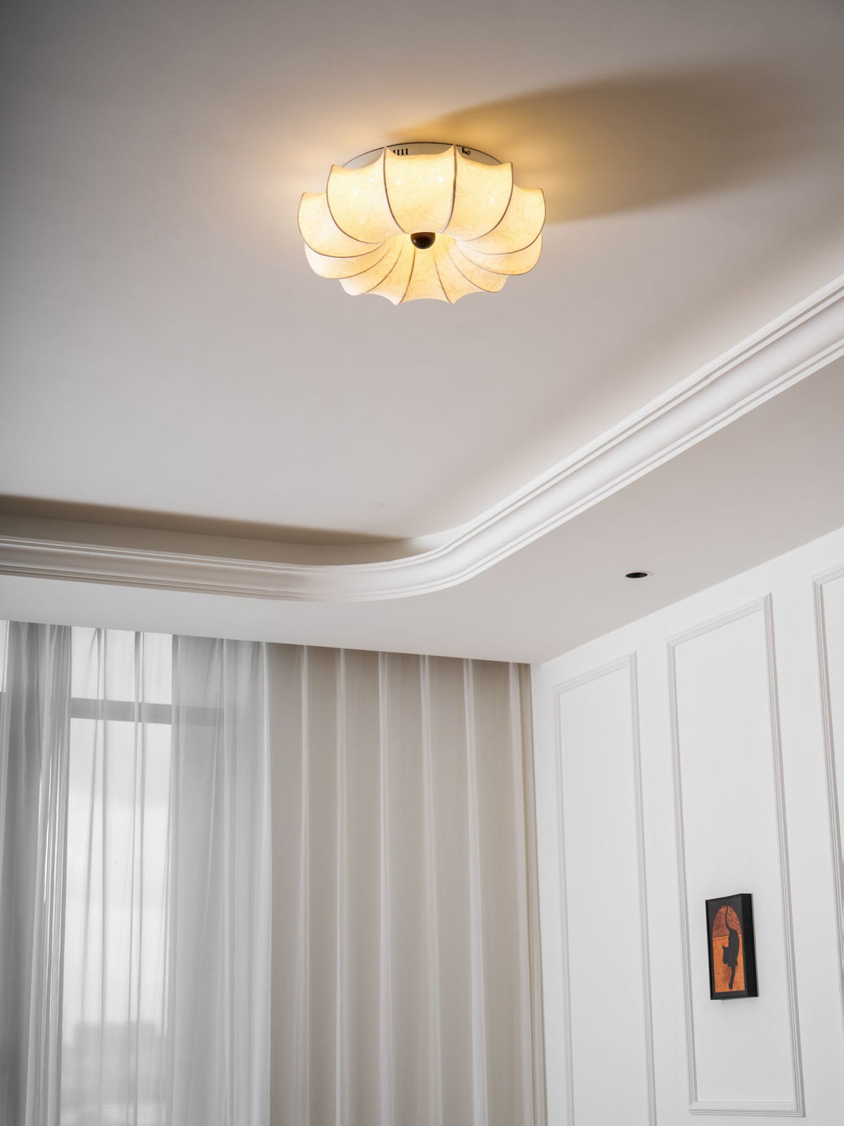 Lit Clérachic ceiling light in a white bedroom with sheer curtains, a coved ceiling edge and a framed print on the wall.