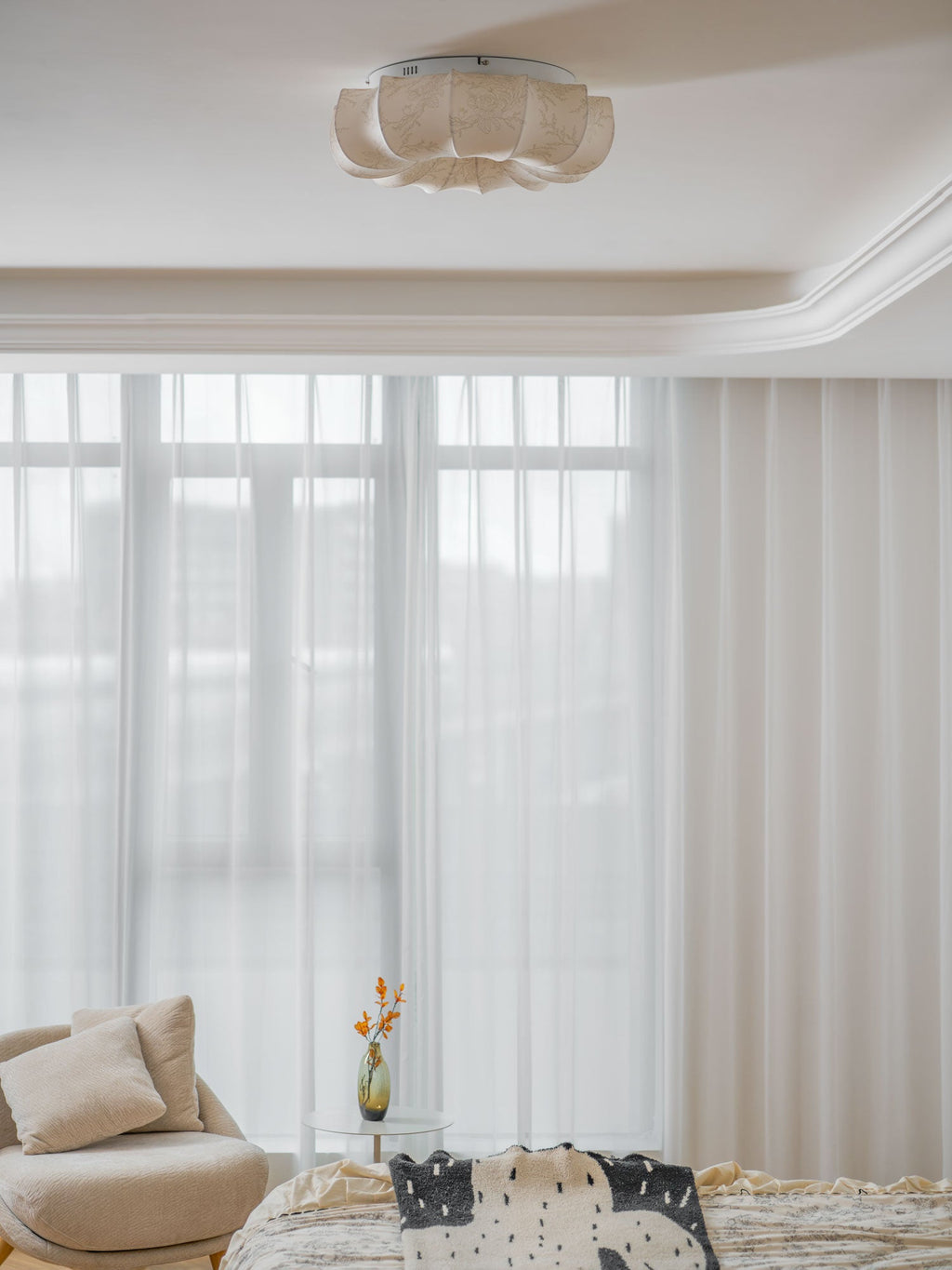 A Clérachic fabric ceiling light, unlit, above a bed with a floral cover and tall sheer curtains.