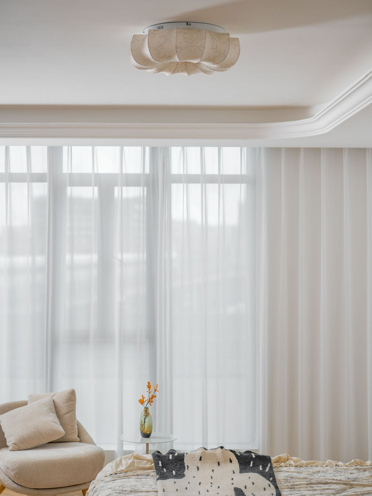 A Clérachic fabric ceiling light, unlit, above a bed with a floral cover and tall sheer curtains.