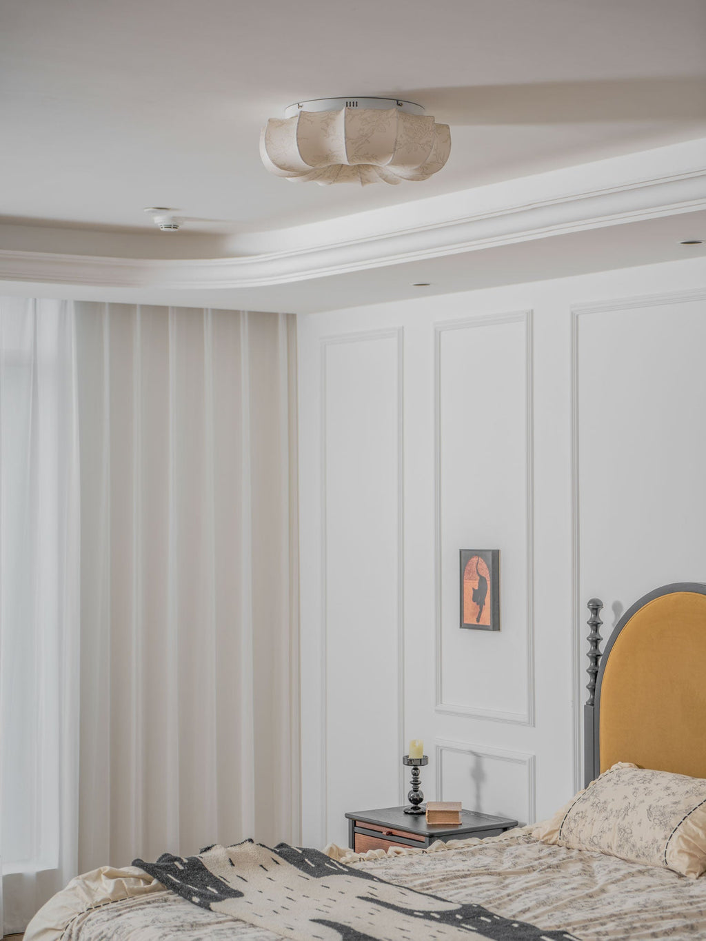 A Clérachic ceiling light, unlit, above a white panelled bedroom with a mustard headboard and a patterned bed cover.