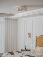 A Clérachic ceiling light, unlit, above a white panelled bedroom with a mustard headboard and a patterned bed cover.