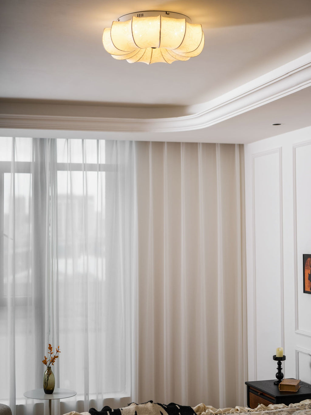 A Clérachic fabric ceiling light glowing in a white room with sheer curtains and a small vase of flowers below.