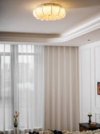 A Clérachic fabric ceiling light glowing in a white room with sheer curtains and a small vase of flowers below.