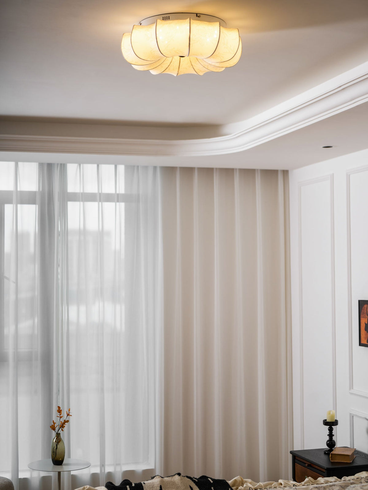 A Clérachic fabric ceiling light glowing in a white room with sheer curtains and a small vase of flowers below.