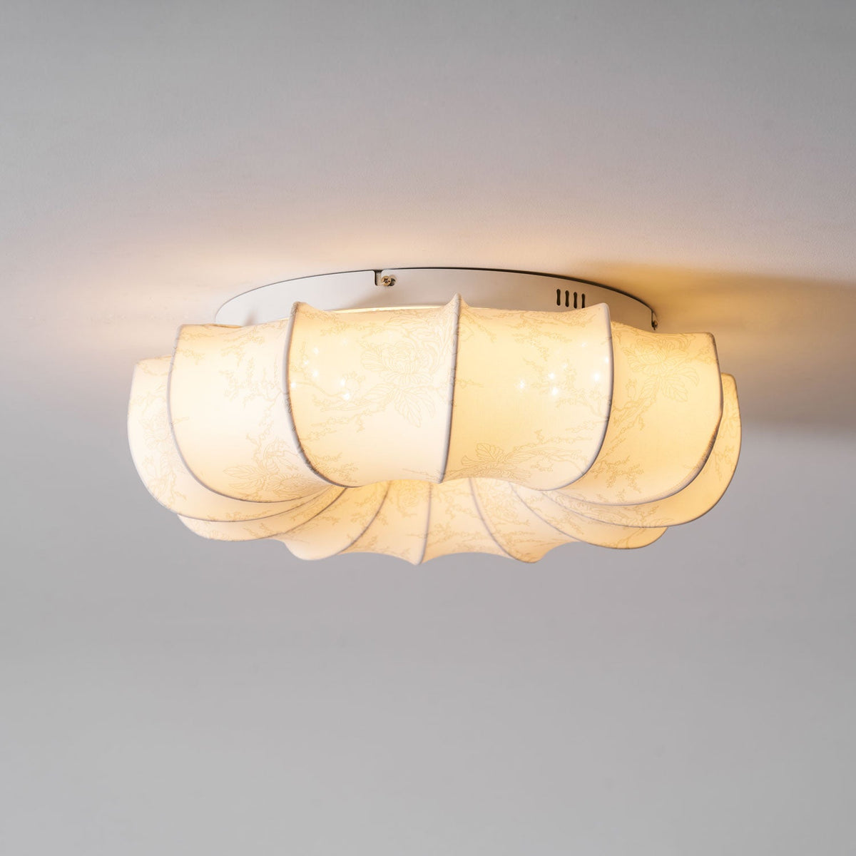 A lit Clérachic fabric ceiling light seen from the side, with warm light spilling onto the ceiling around its edge.