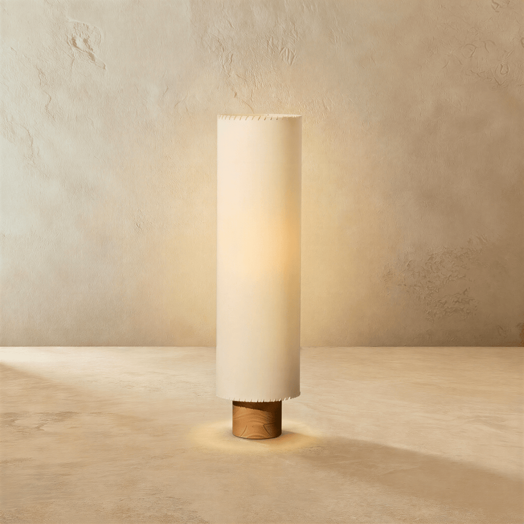 Clérachic floor lamp with a tall beige fabric cylinder shade glowing warm on a short wooden base, against limewashed plaster.
