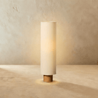 Clérachic floor lamp with a tall beige fabric cylinder shade glowing warm on a short wooden base, against limewashed plaster.