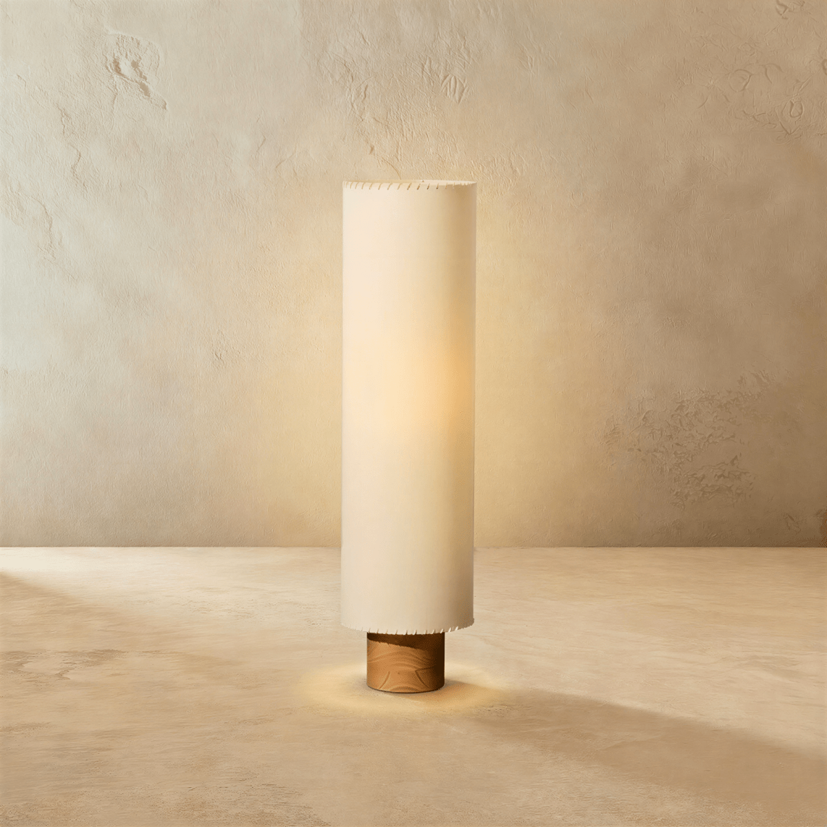 Clérachic floor lamp with a tall beige fabric cylinder shade glowing warm on a short wooden base, against limewashed plaster.
