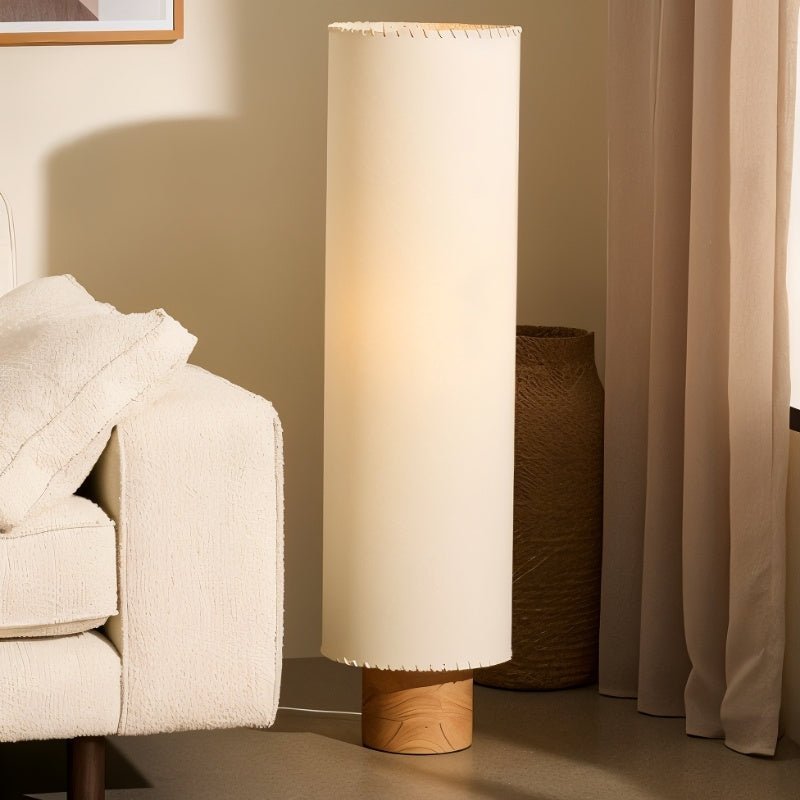 Clérachic floor lamp with a lit beige cylinder shade beside a cream sofa, a woven basket and a pleated curtain.