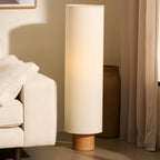Clérachic floor lamp with a lit beige cylinder shade beside a cream sofa, a woven basket and a pleated curtain.