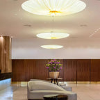 Three Clérachic fabric pendants of different widths glow in a lobby, with a walnut wall, a vase of flowers and a curved chair.