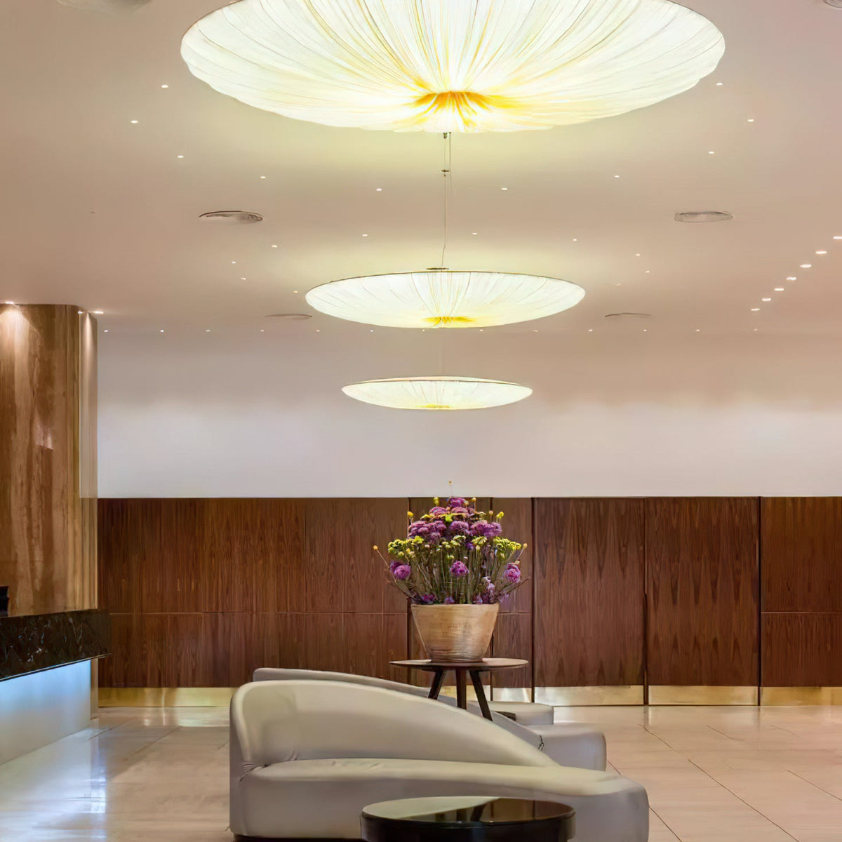 Three Clérachic fabric pendants of different widths glow in a lobby, with a walnut wall, a vase of flowers and a curved chair.