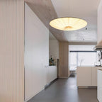 A beige Clérachic fabric disc pendant hangs in a hallway of pale wood panelling, with a kitchen island visible beyond it.
