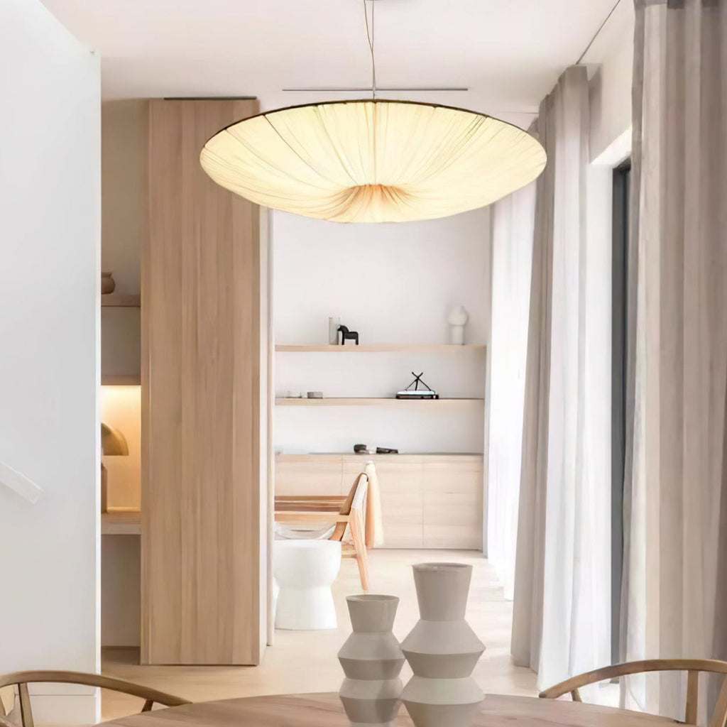 A glowing Clérachic pendant light hangs in a bright open room with pale wood, tall curtains and two white vases below.
