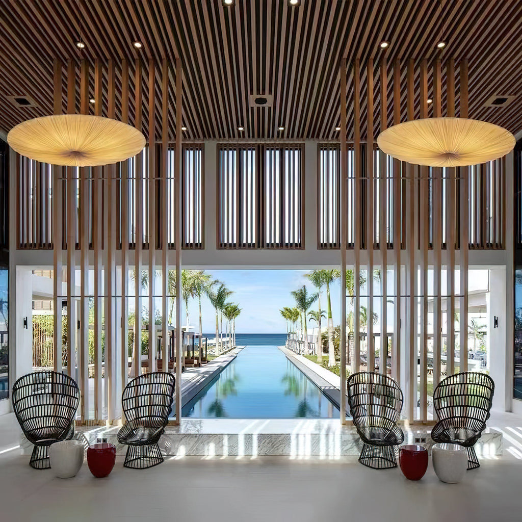 Two large Clérachic fabric pendants hang in a high slatted hall that opens onto a long pool, with black wicker chairs below.