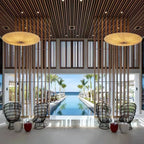 Two large Clérachic fabric pendants hang in a high slatted hall that opens onto a long pool, with black wicker chairs below.