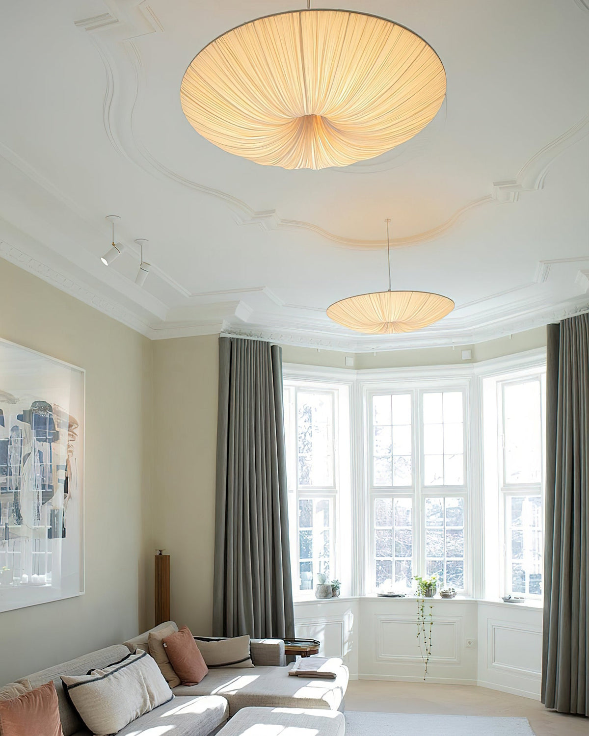Two Clérachic fabric pendants of different sizes hang from a moulded ceiling in a room with a bay window and a pale sofa.