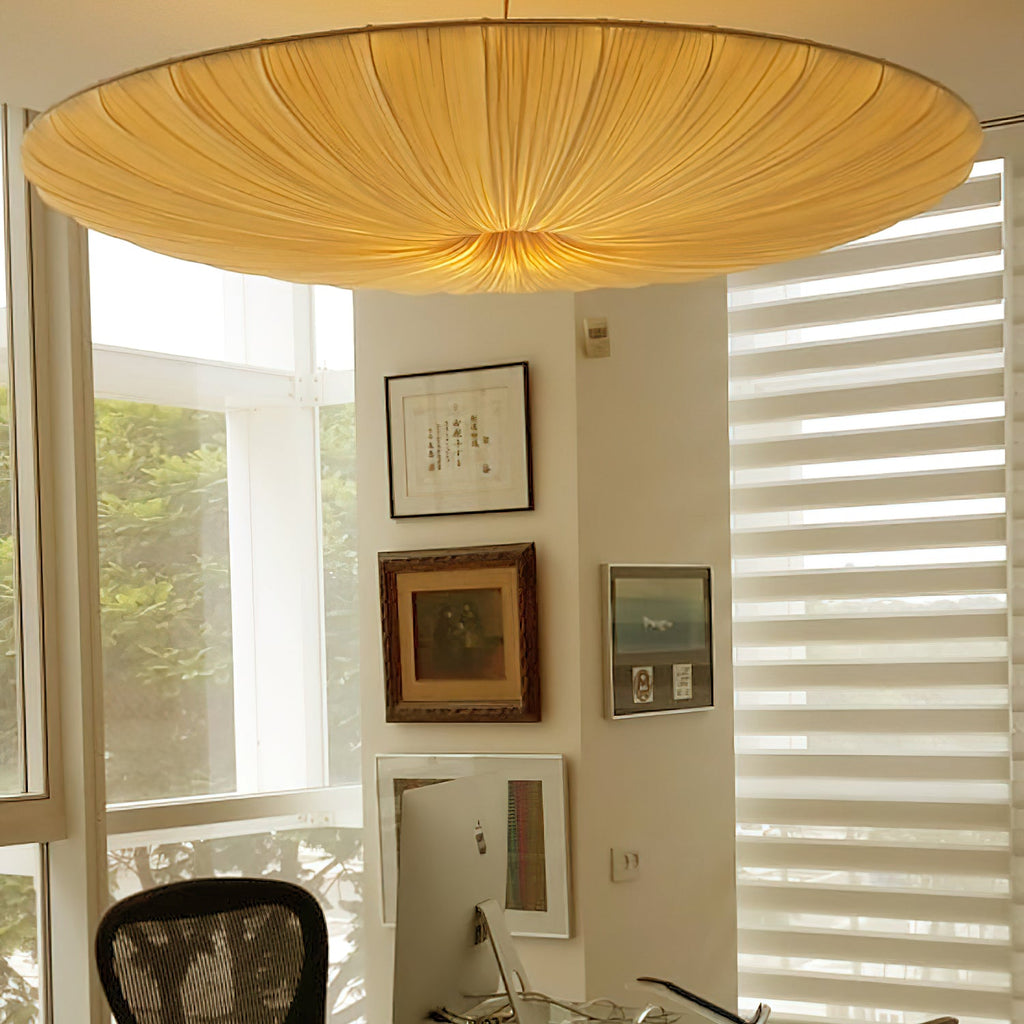 Close view of the pleated beige fabric of this Clérachic pendant, glowing warm above a study with framed art and a window.