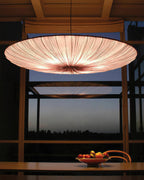 A Clérachic pendant with a pink-tinted glow hangs over a wooden table with a bowl of fruit, in front of a dusk view through large windows.