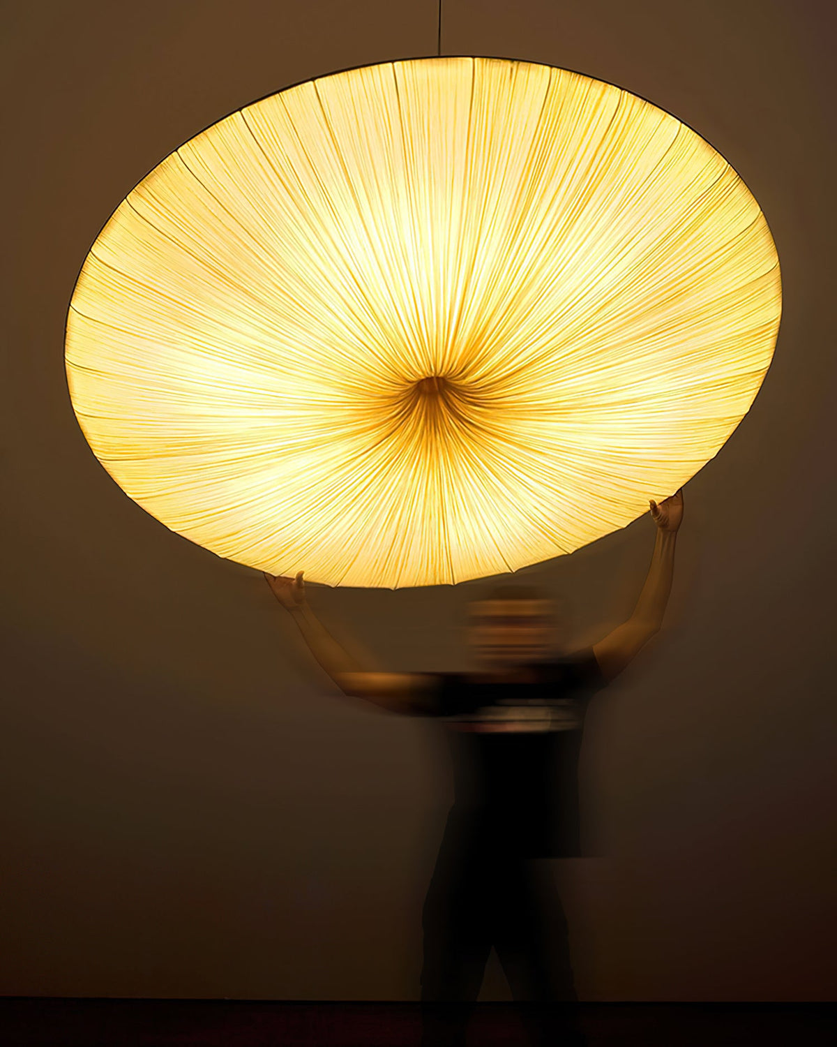 The underside of this Clérachic pendant glows amber, its gathered fabric radiating from the centre, with a blurred figure holding it up.