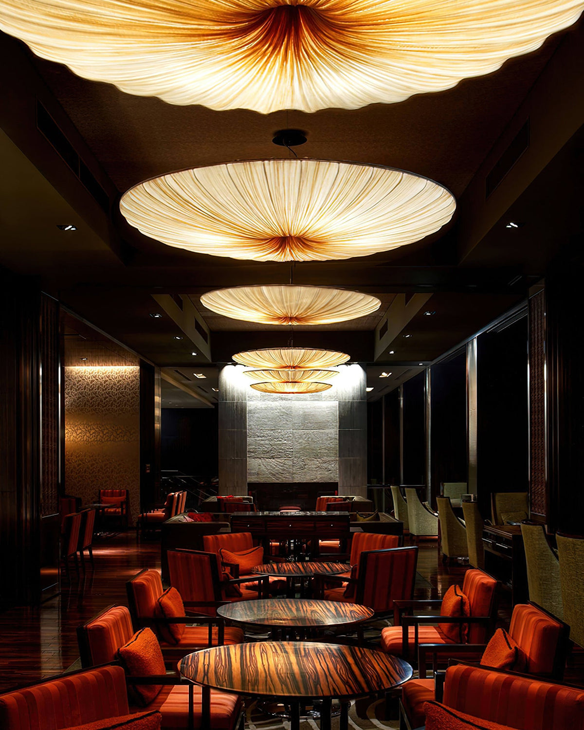 Several Clérachic fabric pendants in a row glow above red-chaired tables in a long, dark restaurant.