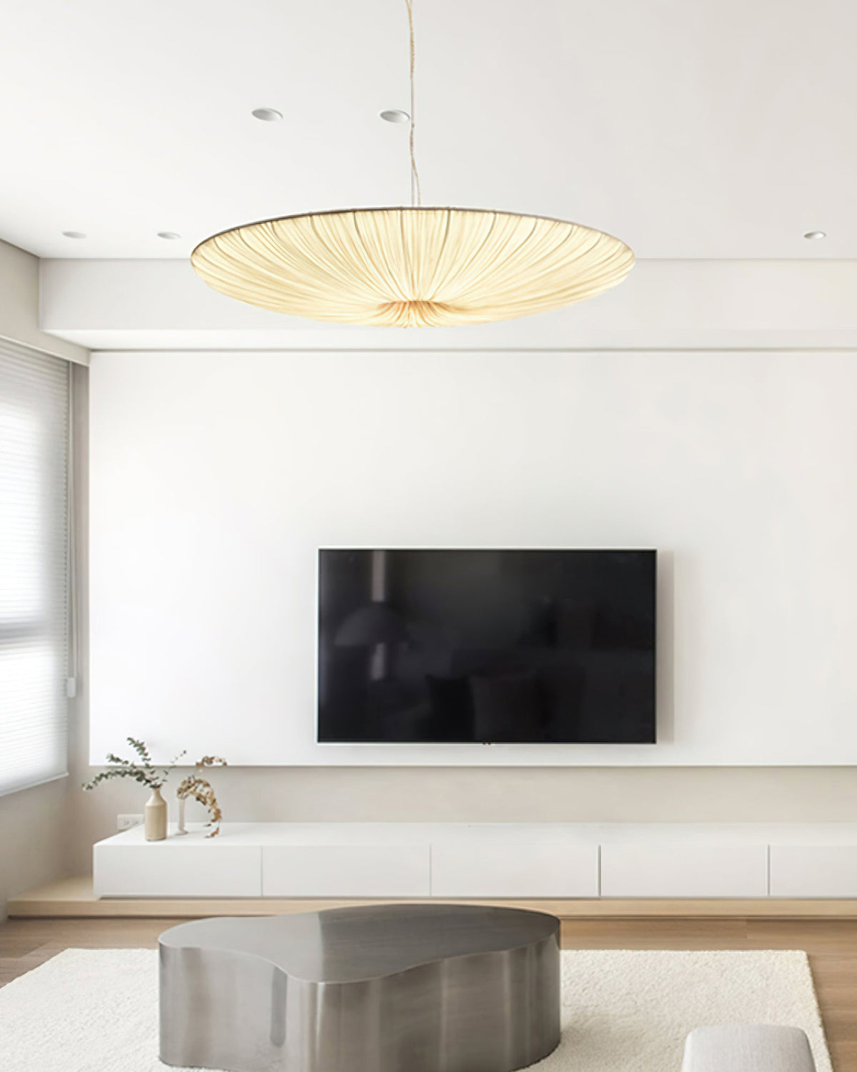 A beige Clérachic pendant light hangs in a white living room, over a low steel coffee table, with a wall-mounted television behind.