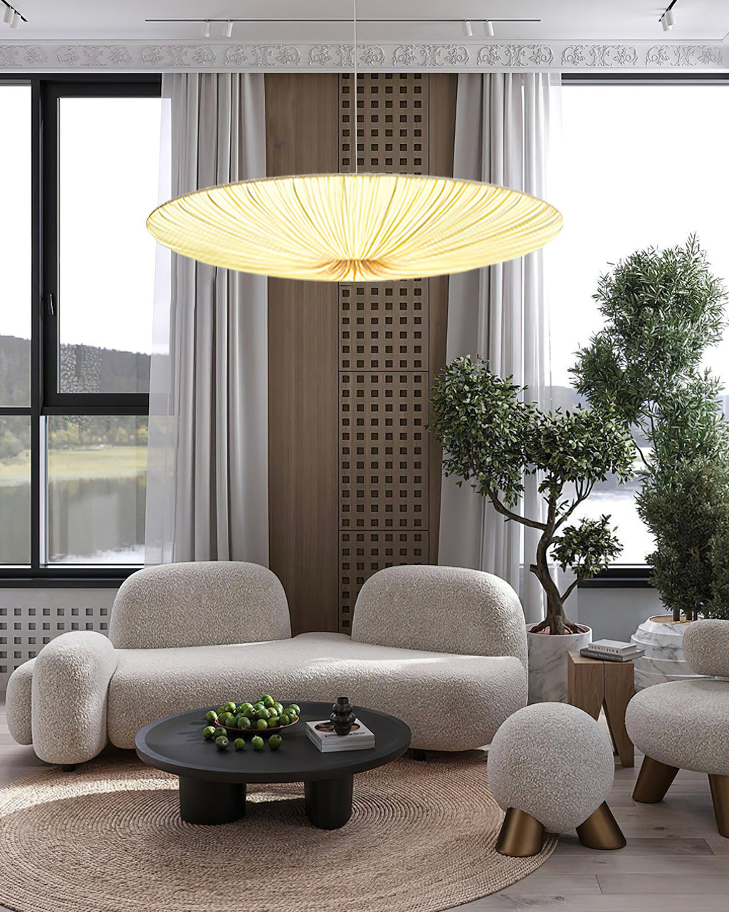 A shallow Clérachic fabric pendant glows over a curved cream sofa and a round coffee table, with tall windows and a potted tree beside.