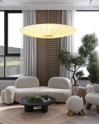 A shallow Clérachic fabric pendant glows over a curved cream sofa and a round coffee table, with tall windows and a potted tree beside.