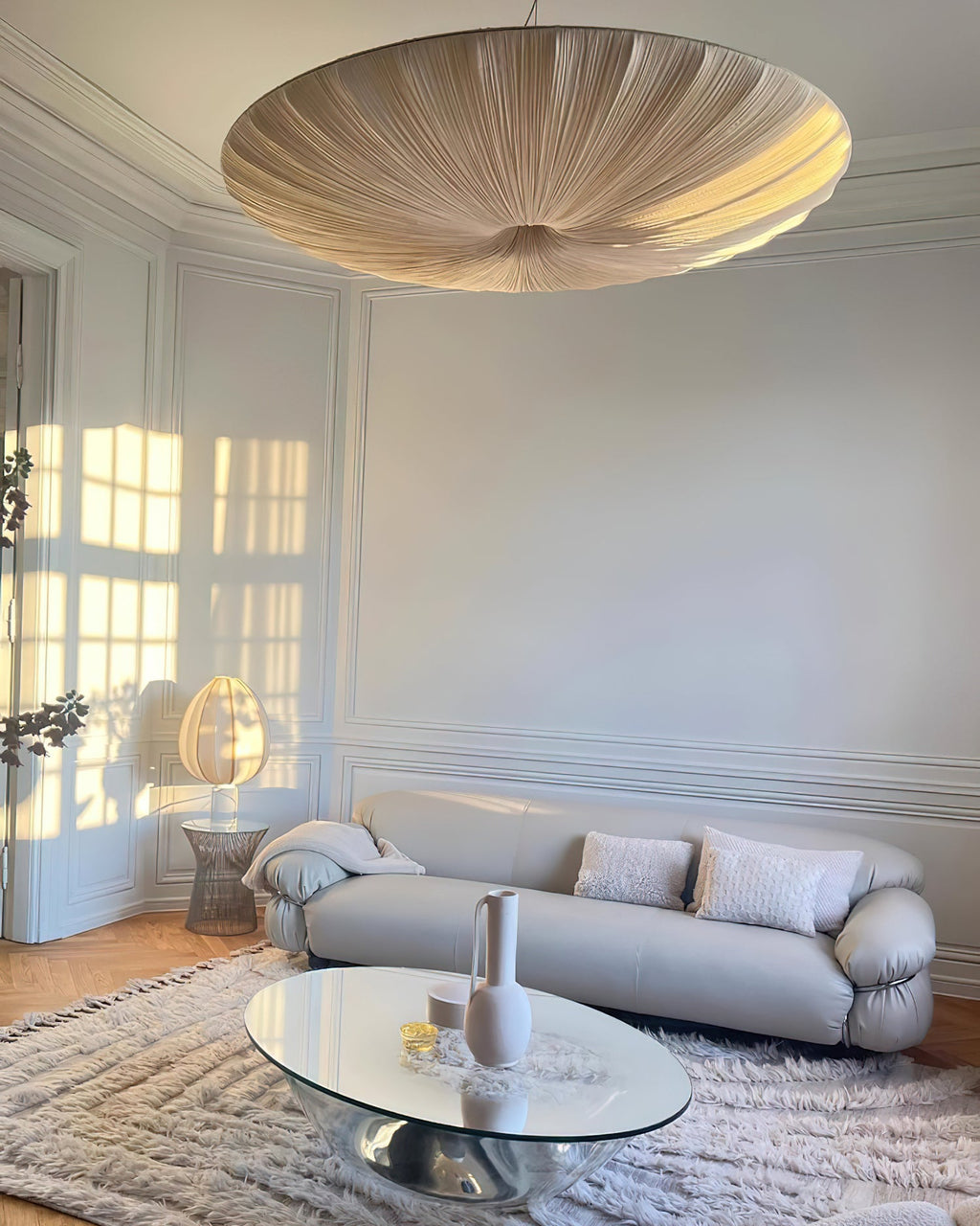 A greyish-beige version of this Clérachic pendant hangs low in a pale panelled room, over a grey sofa and a glass coffee table.