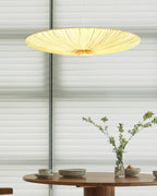 A glowing Clérachic fabric pendant hangs above a round wooden table set with plates and a vase of flowering stems, before blinds.