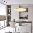 A beige Clérachic pendant hangs over a long grey desk with white chairs, a built-in shelf unit and a window with a pleated blind.