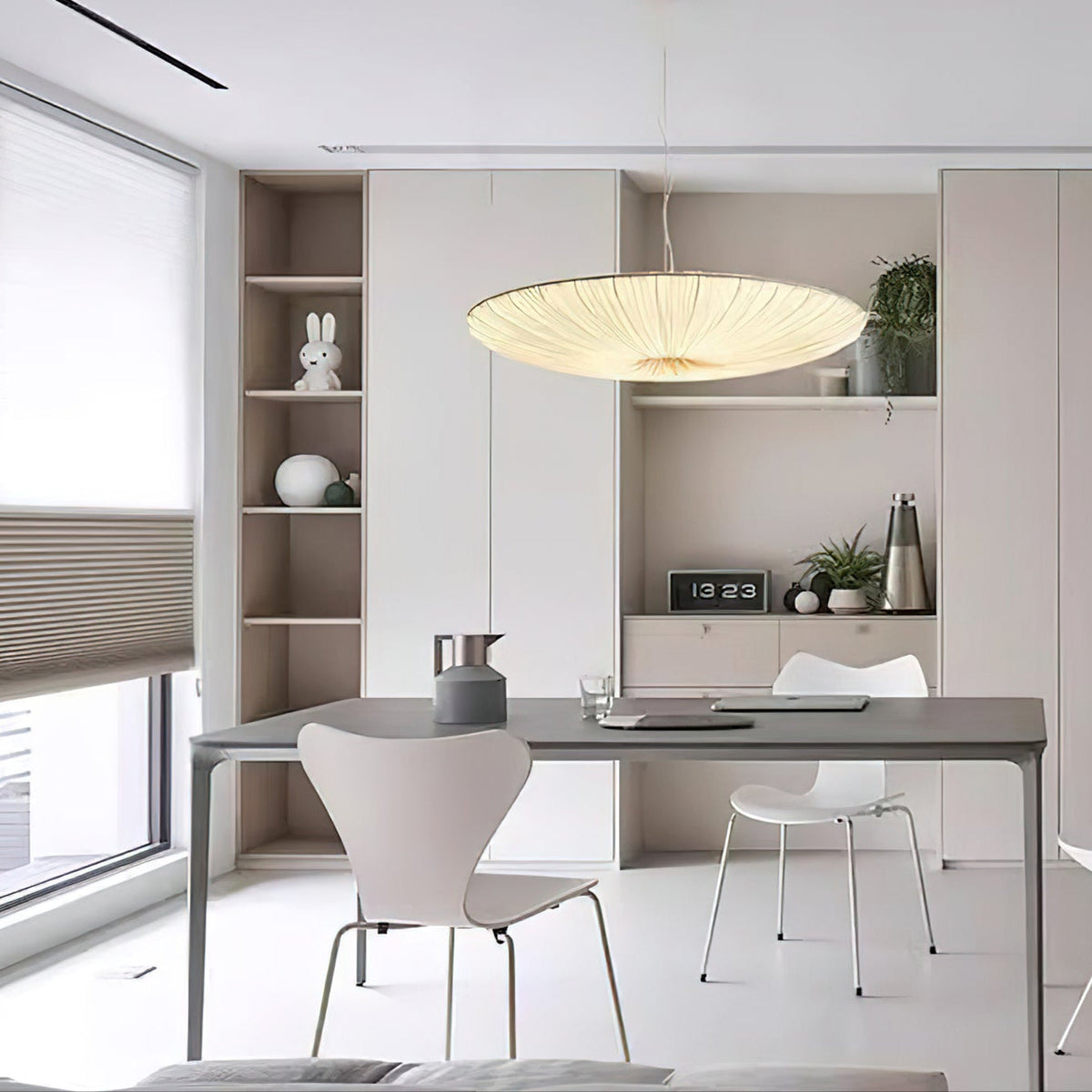 A beige Clérachic pendant hangs over a long grey desk with white chairs, a built-in shelf unit and a window with a pleated blind.