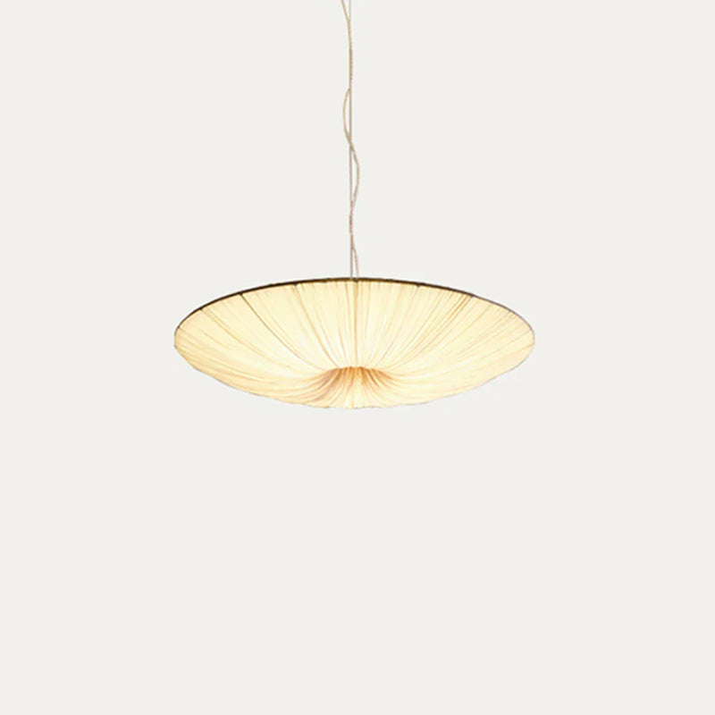 A shallow beige Clérachic fabric pendant lit warm, with its gathered folds meeting at the centre, against a soft grey backdrop.