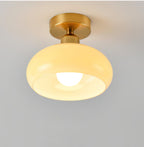 A lit Clérachic ceiling light with a beige glass dome and brass canopy, the round bulb glowing inside, on a grey backdrop.