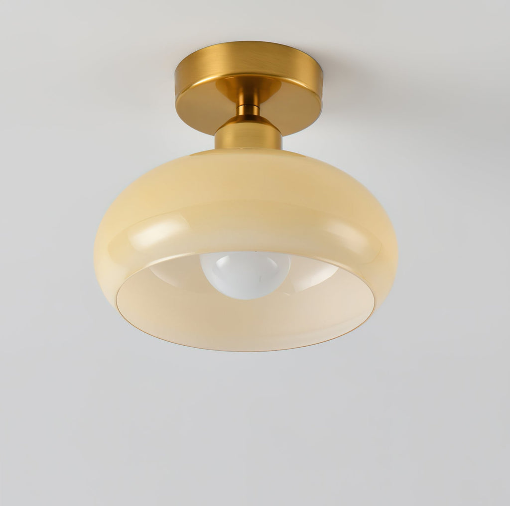 A Clérachic ceiling light with a pale beige glass dome and brass canopy, seen from slightly below against a light wall.