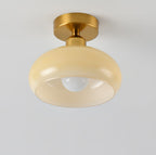 A Clérachic ceiling light with a pale beige glass dome and brass canopy, seen from slightly below against a light wall.